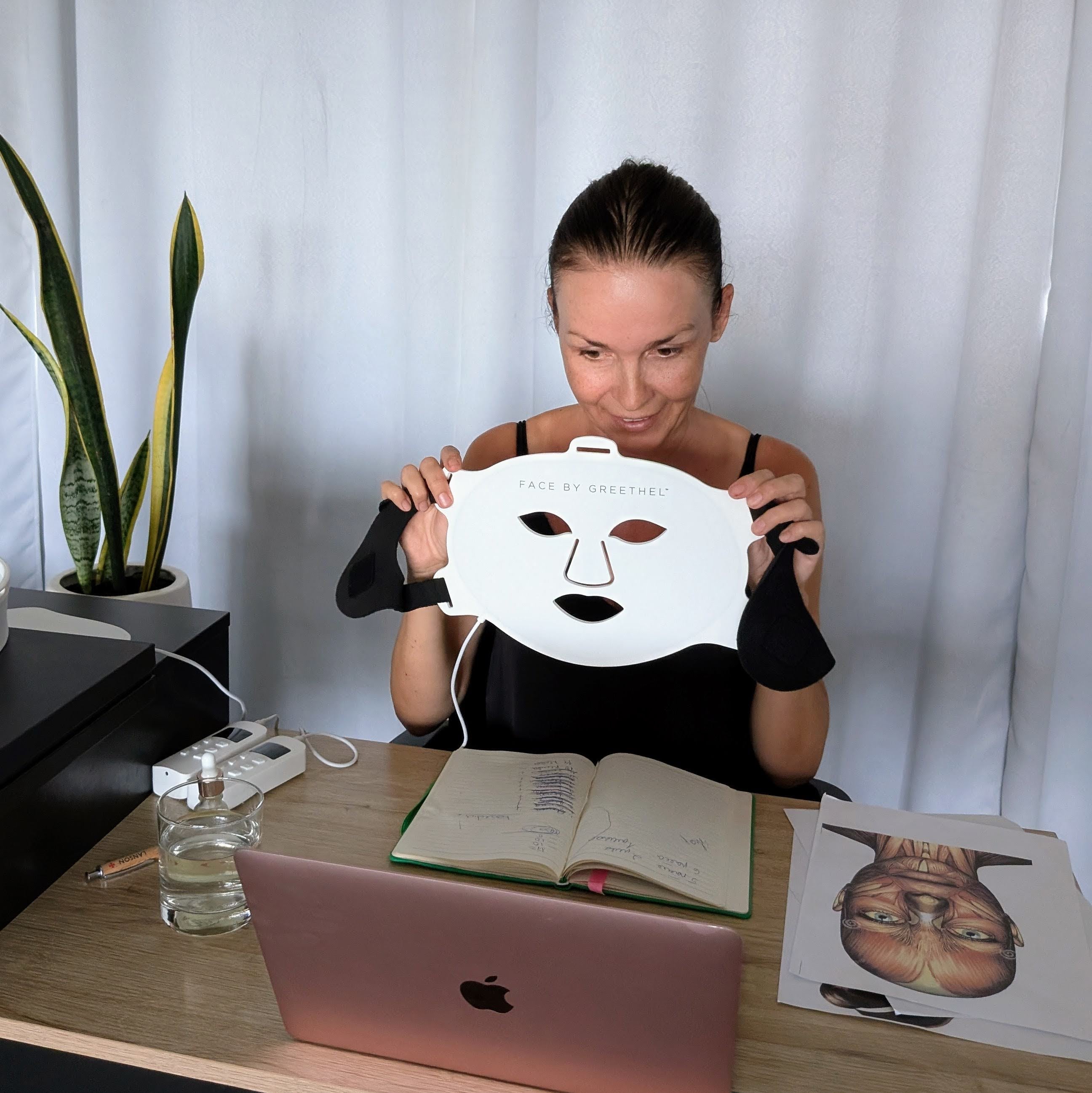 Person holding a face mask in front of a laptop with a plant and books in the background