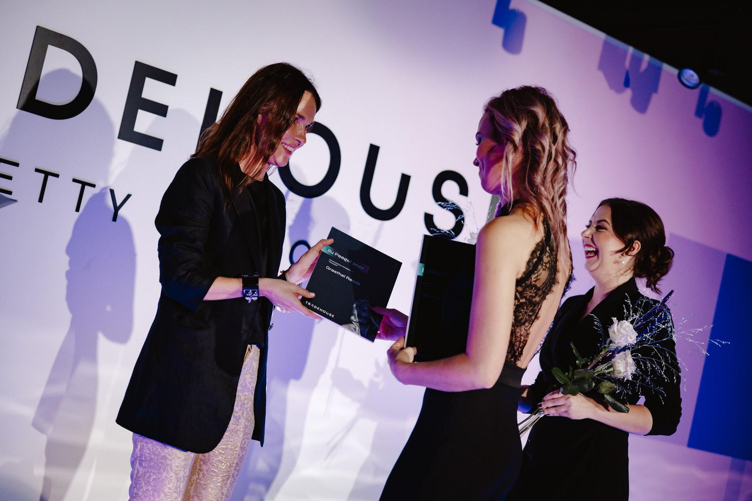 Two women receiving an award on a stage with a visible brand logo in the background.